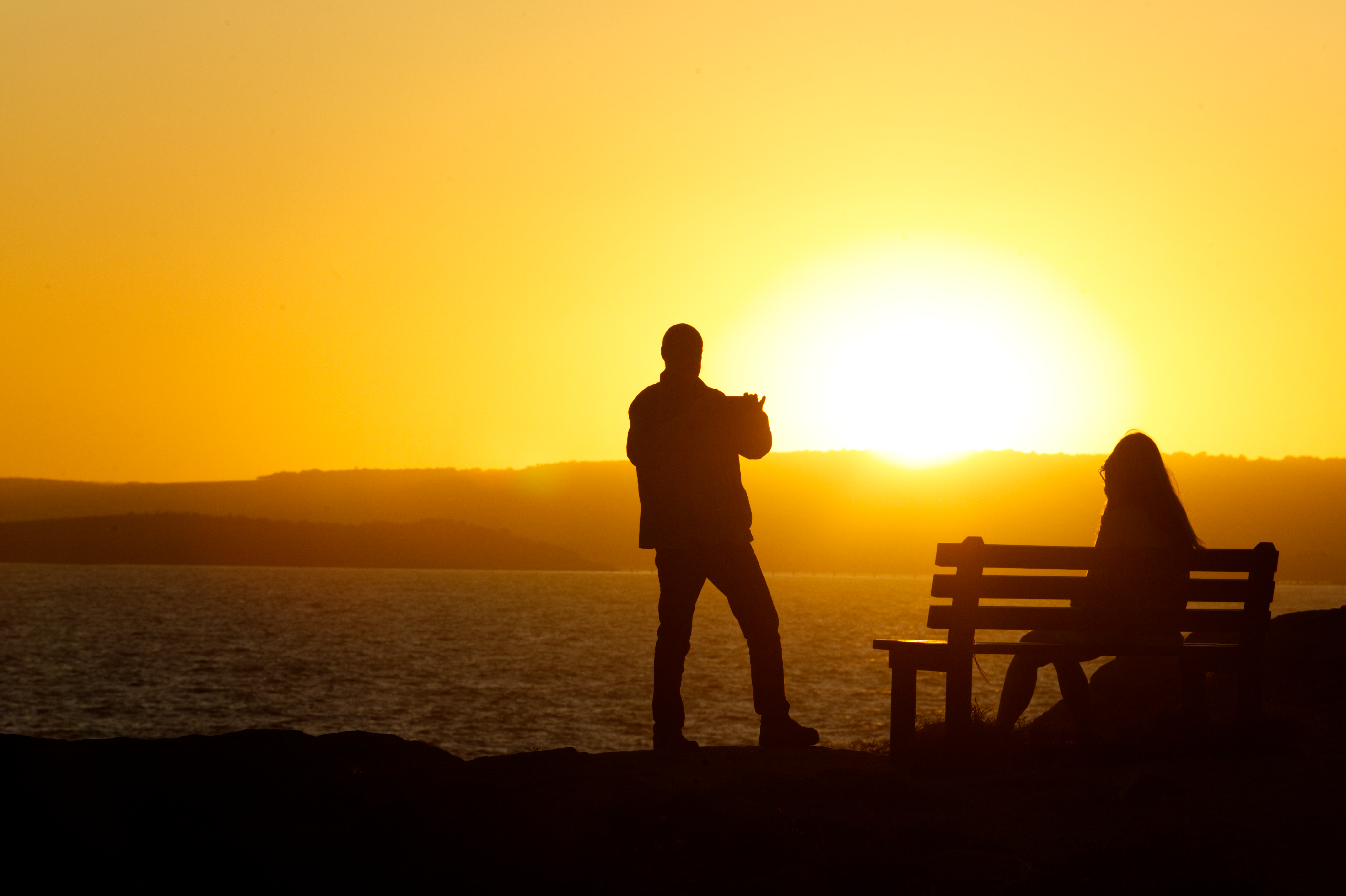 Silhouette with golden setting sun of man taking a photo of a woman on a park bench.