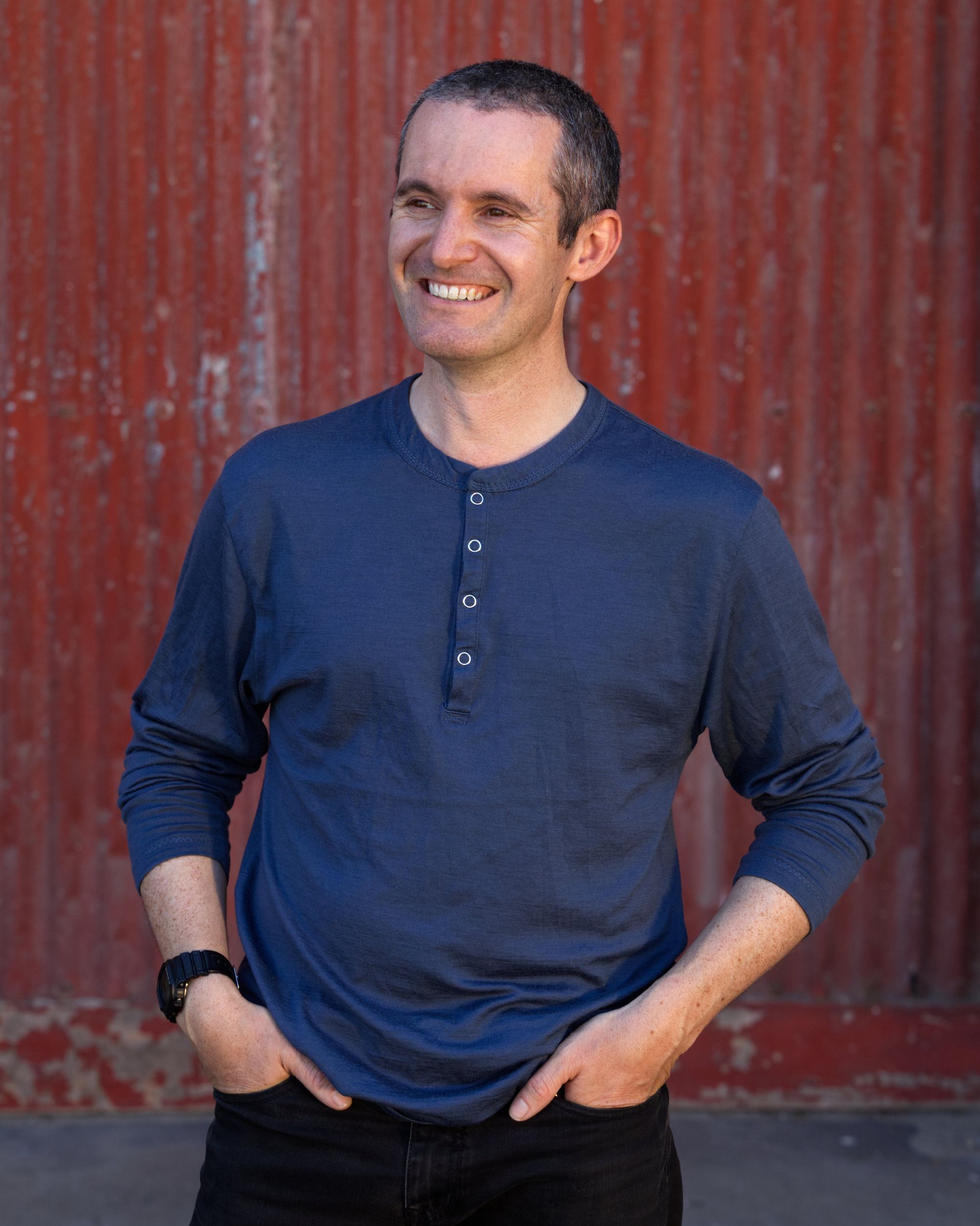 A man in a blue long sleeve tee stands with his hands in his pockets (thumbs out) in front of a red corrugated iron door.