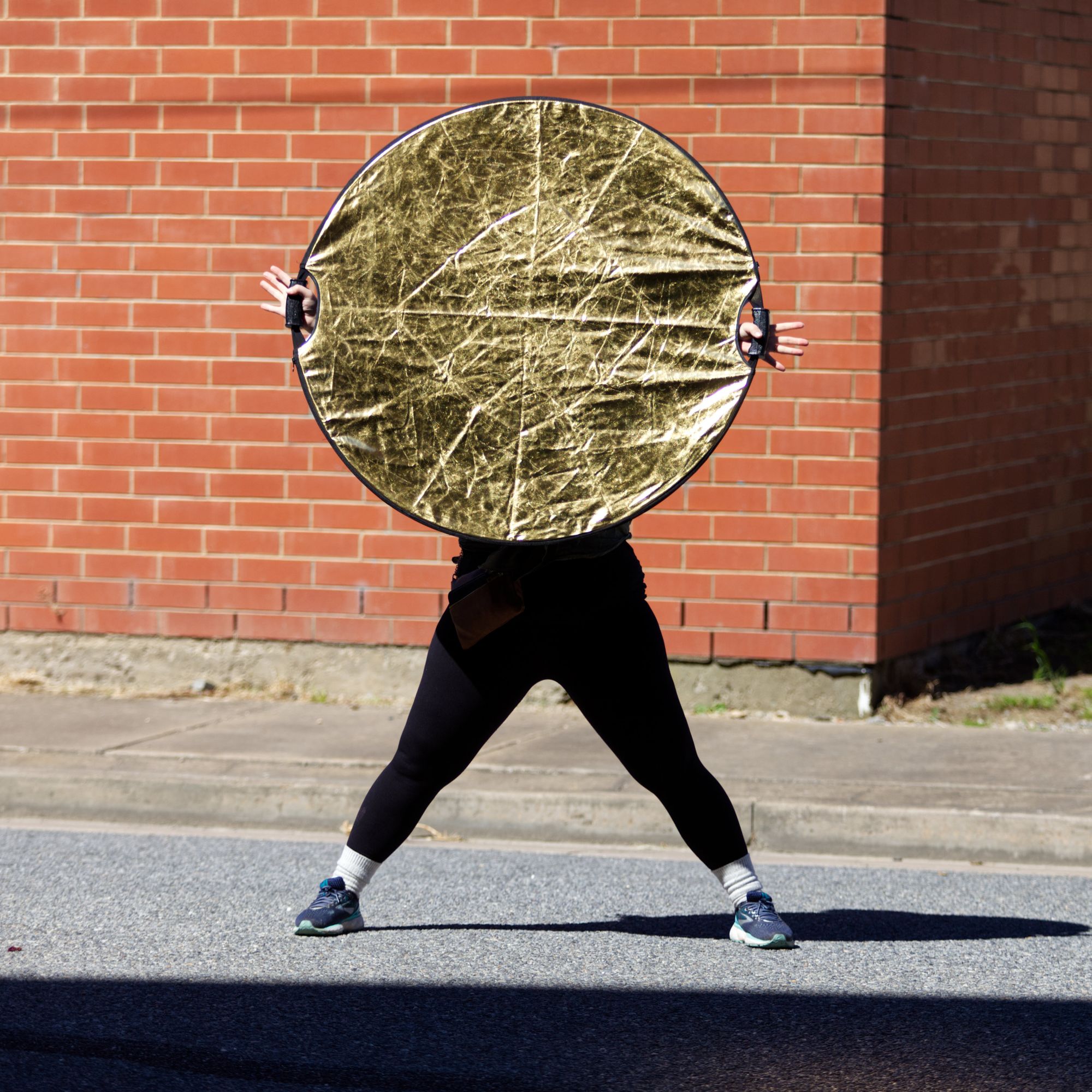 A woman in black leggings obscures her torso and head with a large circular golden reflector.