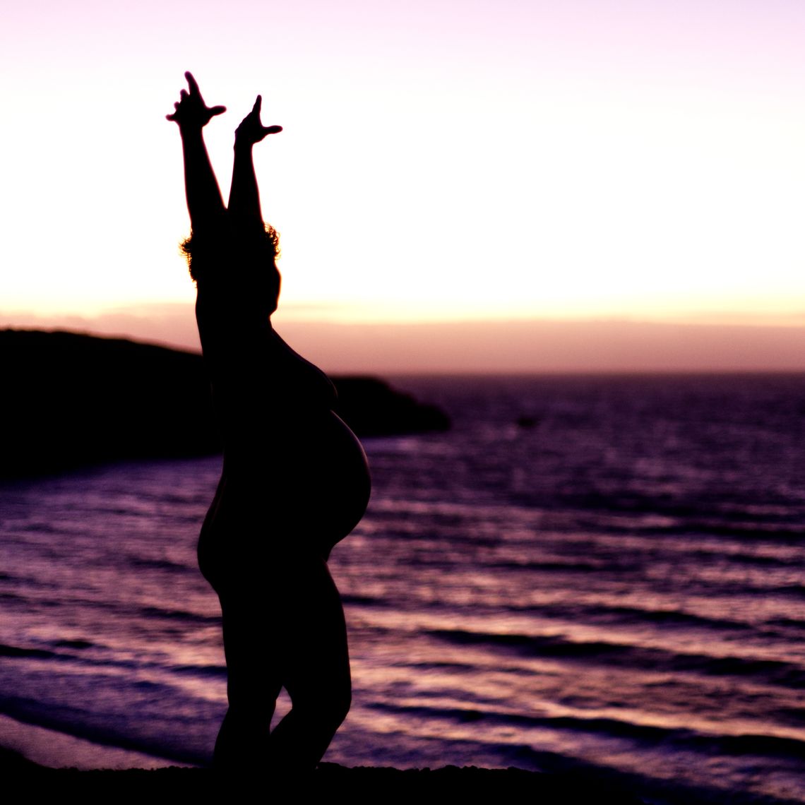 Silhouette of a naked preganant woman against a glowing sky and the ocean.