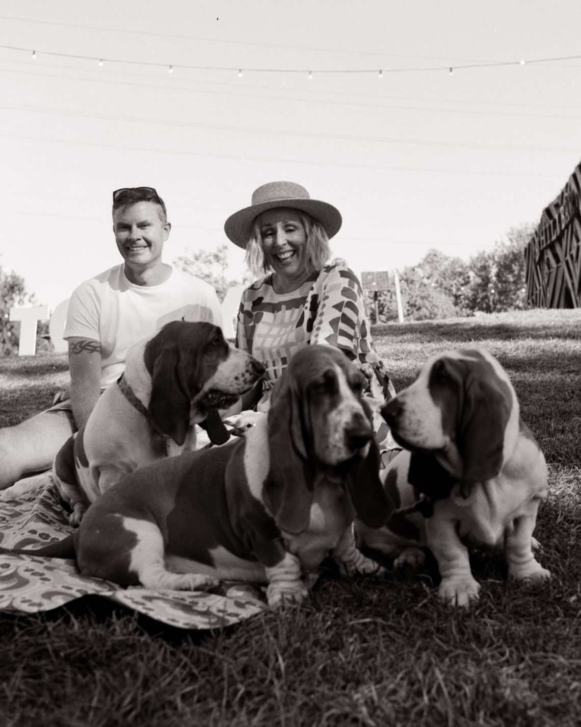 A man and a woman are seated on a rug behind three bassest hounds.