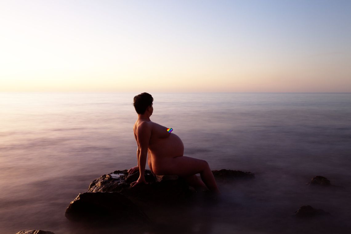 A nude pregnant woman sits on a rock in a blurry ocean.