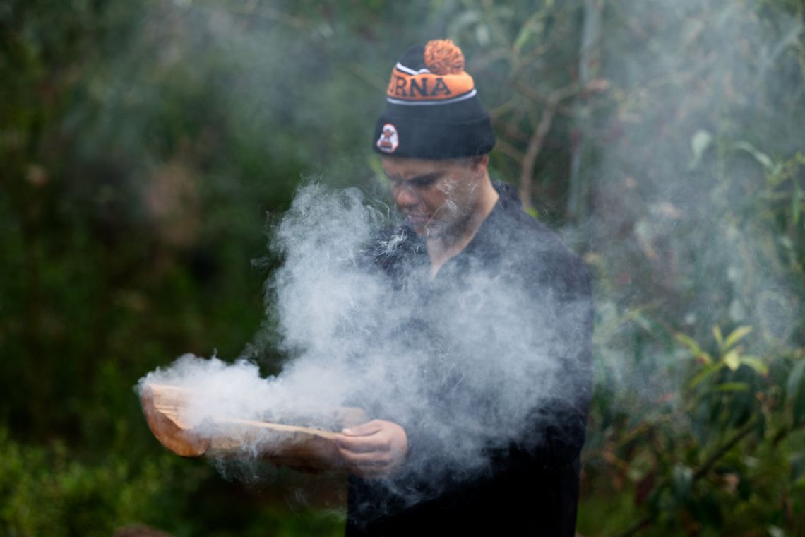 A man in a beanie holds a piece of wood with a lot of smoke in the scene.