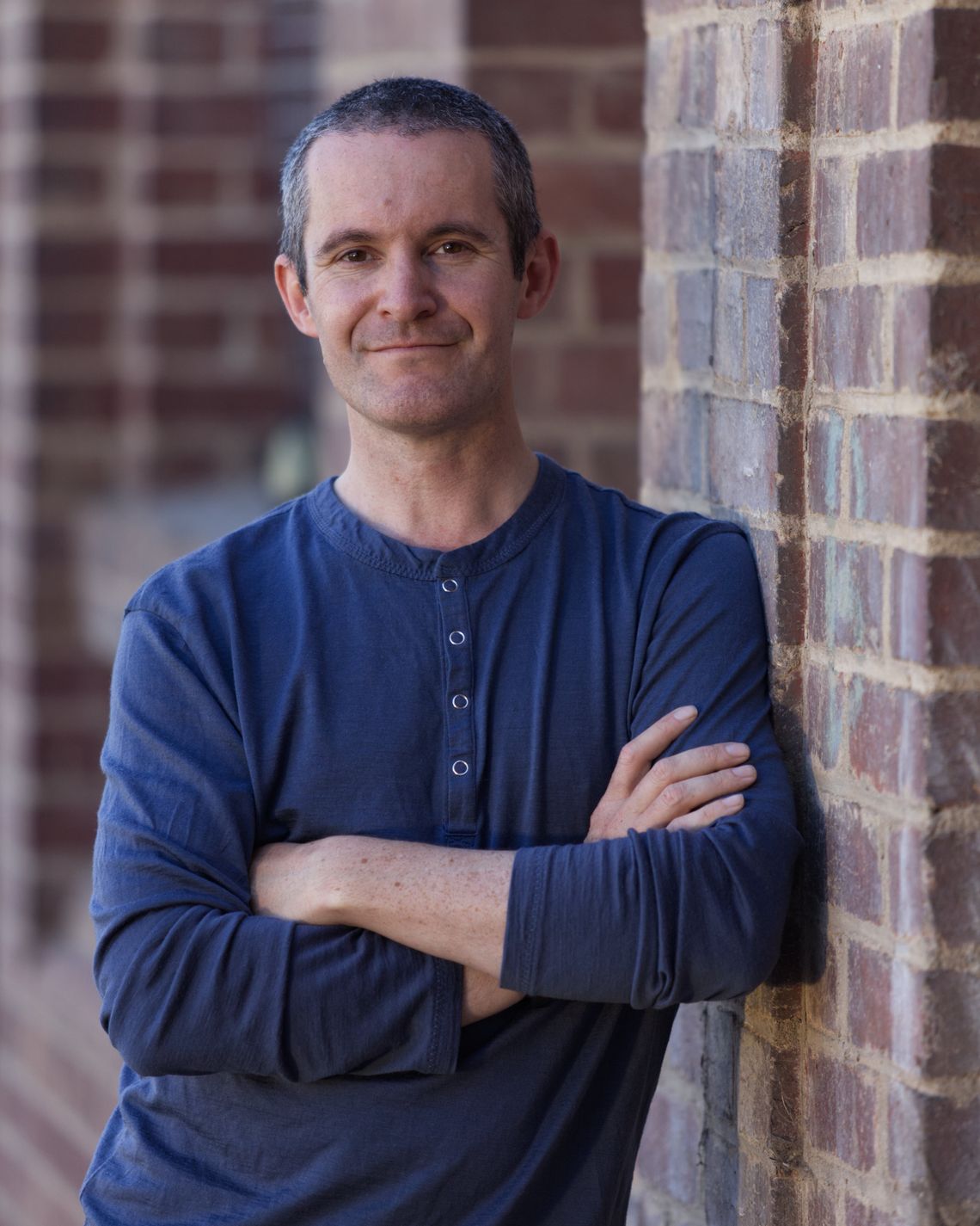 A man in a blue long sleeved tee leans up against a red-brick wall.