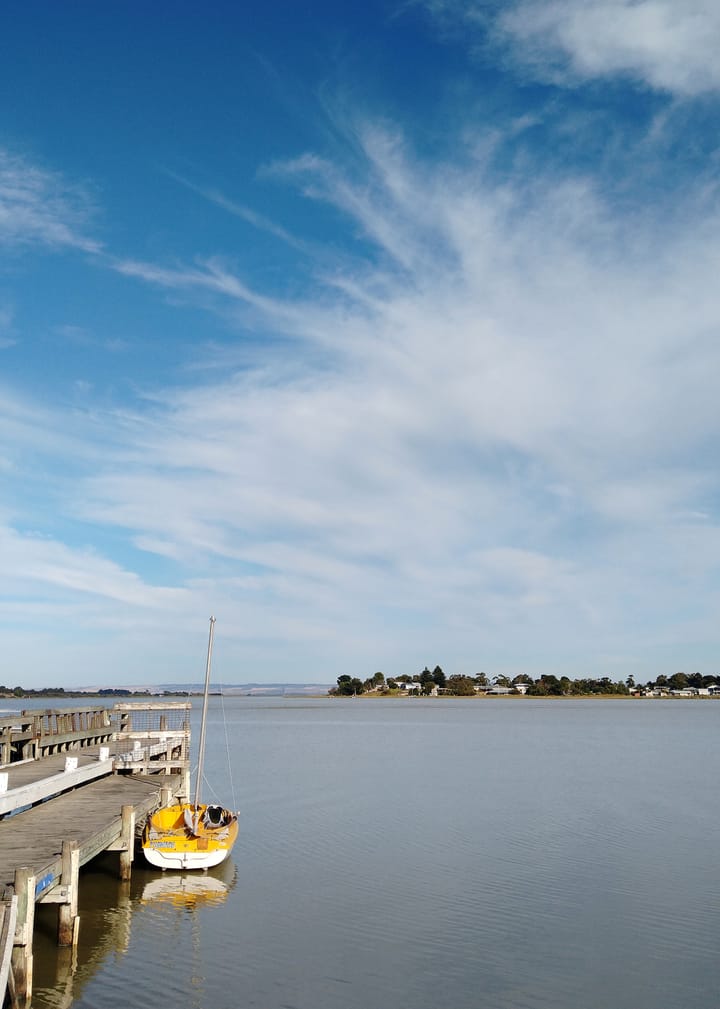 A yellow and white boat tied to a wooden jetty on the side ofthe image. A lake and clouds in the background.